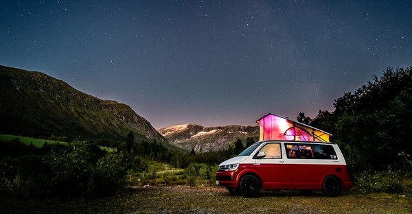 Roter Campervan unter sternenklarem Himmel in einer Berglandschaft mit buntem Licht im Inneren.