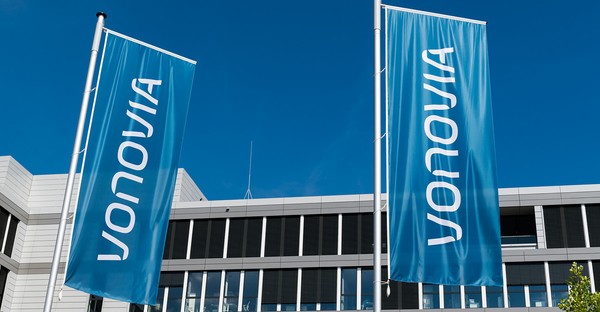 Zwei blaue Vonovia-Flaggen wehen vor einem modernen Gebäude unter klarem Himmel.