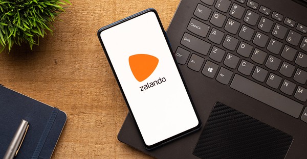 Smartphone mit Zalando-Logo liegt auf einem Laptop, daneben ein Notizbuch und eine Pflanze.