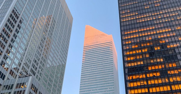 Drei Wolkenkratzer bei Sonnenuntergang, der mittlere leuchtet rosa, die anderen reflektieren Licht.