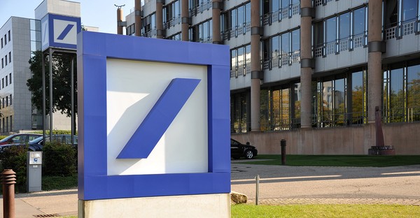 Blaues Logo vor einem modernen Bürogebäude mit Glasfassade und parkenden Autos.