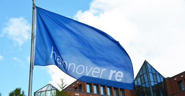 Blaue Flagge mit der Aufschrift "Hannover Re" weht vor einem Gebäude mit Glasdach.