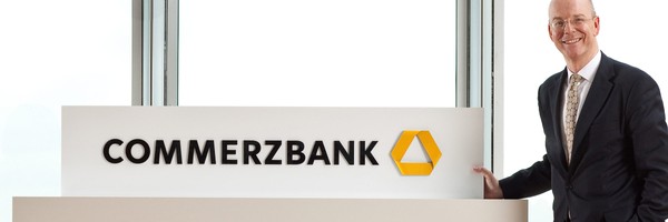 Mann im Anzug steht neben einem Commerzbank-Schild mit Logo.