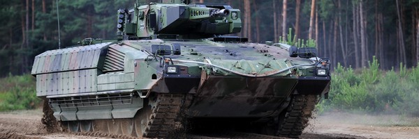 Ein grüner Panzer fährt durch einen Wald auf einem sandigen Weg.