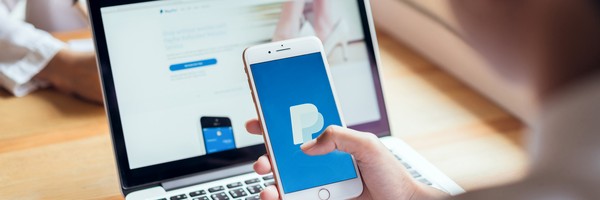 Person hält ein Smartphone mit PayPal-Logo vor einem Laptop mit geöffneter PayPal-Website.